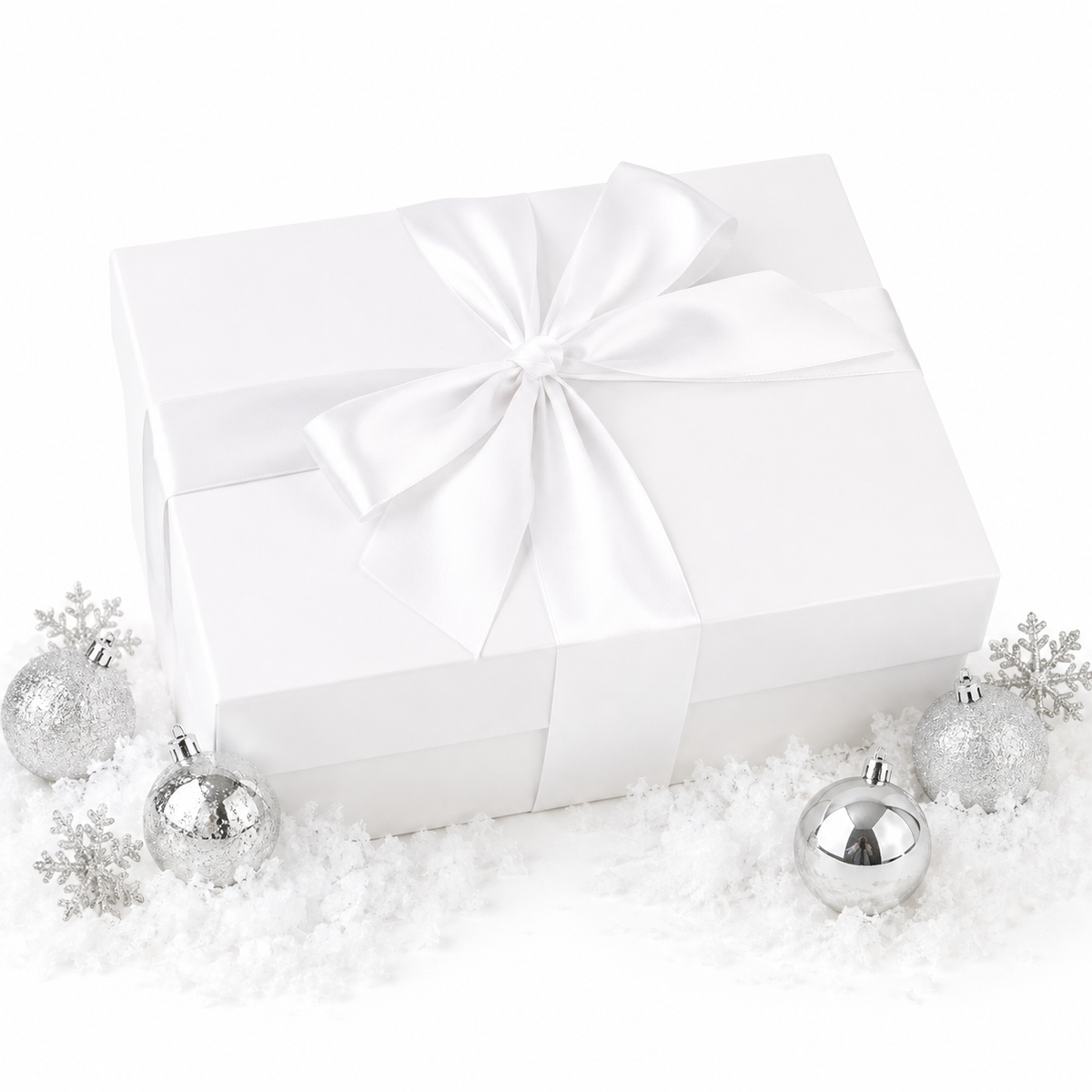 A white gift box wrapped with a large white satin ribbon and bow, styled on a white surface with scattered faux snow and silver holiday ornaments.
