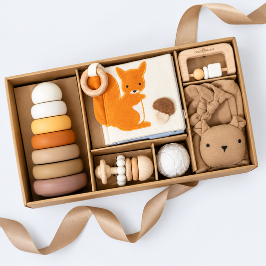A wooden and fabric baby gift set arranged in a kraft cardboard box with ribbon, featuring a colorful ring stack, a fox-themed cloth book, a wooden teething toy, a plush bear, and other soft baby accessories on a white background.