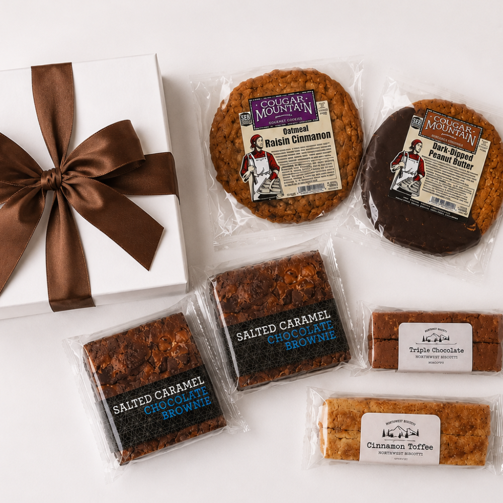 A gift assortment of cookies and brownies from Cookie Monster's Mountain, including packaged salted caramel chocolate brownie, salted caramel chocolate cookie, salted cinnamon cinnamon, and triple chocolate cookie, arranged on a white surface next to a white gift box with a brown ribbon.