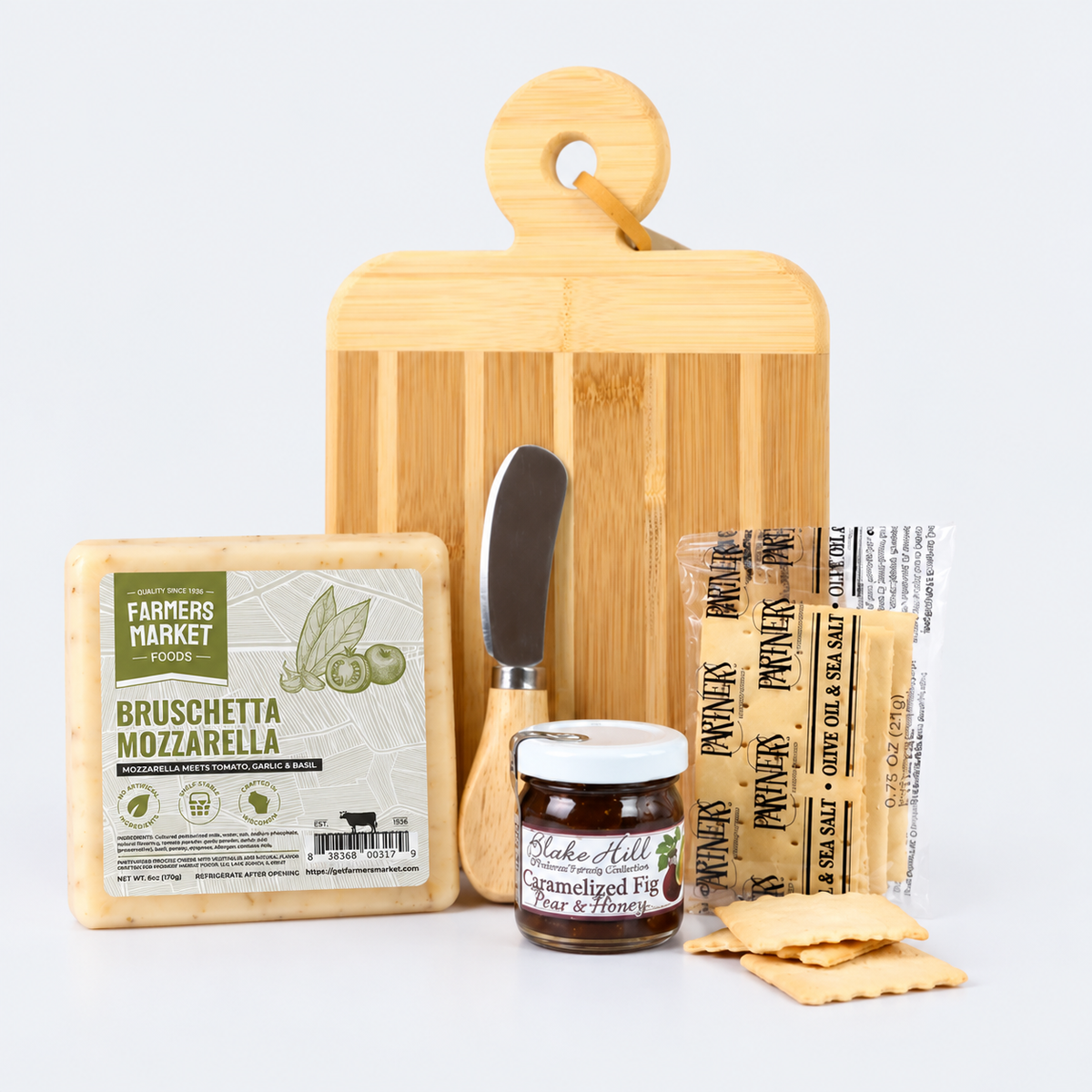 A gourmet cheese and crackers gift set arranged on a light gray background, featuring a wedge of Parmigiano d'Orino Italian cheese, a bamboo cutting board, a small cheese knife, a jar of prosciutto crudo, packaged prosciutto, and flatbread crackers.