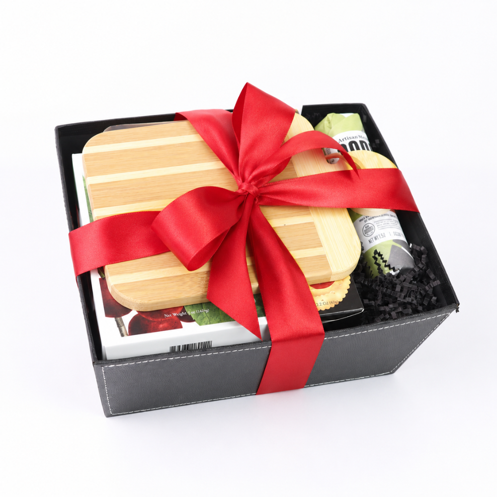 A black gift box containing a bamboo cutting board, a red satin ribbon, and a small packaged item, photographed on a white background.
