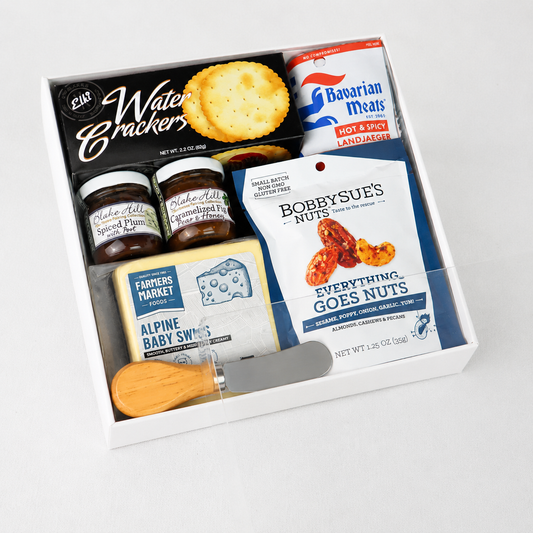 A curated gourmet gift box containing a variety of food items, including Bobbysue's Everything Goes Nuts, Alpine Baby Snacks, Bavarian Meats, and Water Crackers, arranged neatly inside a white box with a small wooden-handled knife.