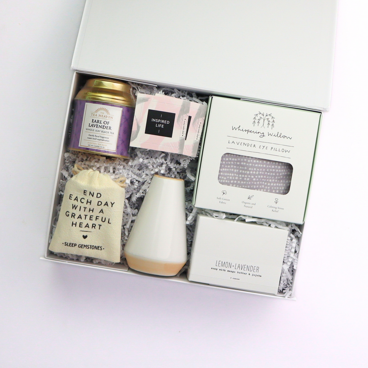 Gift set with wellness products including a tea canister, eye pillow, and sleep stones on a white background.  Top-down view of Lavender Self-Care Gift Box featuring a tea tin, soap bar, lavender sachet, and ceramic bud vase